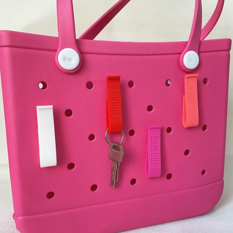 K&ouml;nnte beinhalten: Eine leuchtend rosa Tragetasche mit wei&szlig;en Griffbefestigungen und einer Reihe kreisf&ouml;rmiger Perforationen. Rote, rosa und orangefarbene Schl&uuml;sselanh&auml;nger sind an der Tasche befestigt, auf denen die Namen "Tiffany", "Browne" und "Susanna" aufgedruckt sind. Ein Schl&uuml;ssel ist am Schl&uuml;sselanh&auml;nger "Tiffany" befestigt.