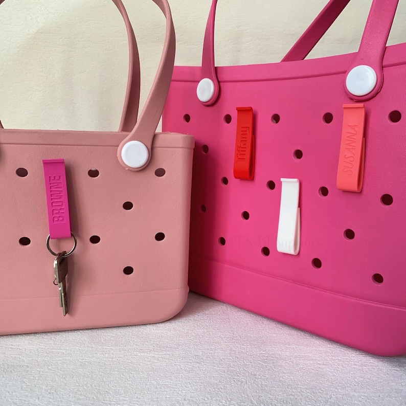 K&ouml;nnte beinhalten: Zwei rosa Tragetaschen. Die kleinere Tasche ist hellrosa mit einem Schl&uuml;sselanh&auml;nger mit der Aufschrift "BROWNE". Die gr&ouml;&szlig;ere Tasche ist pink mit Anh&auml;ngern mit der Aufschrift "Tiffany" und "SUSANNA". Beide Taschen haben wei&szlig;e Griffbefestigungen.