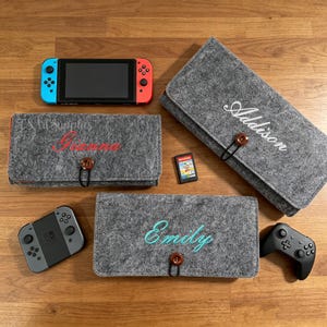 May include: A collection of gray felt cases with embroidered names, designed for a gaming console. The cases feature a button closure and are displayed with a console, controllers, and a game cartridge. The names embroidered are "Addison", "Gianna", and "Emily".