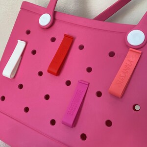 K&ouml;nnte beinhalten: Eine leuchtend rosa Tragetasche mit wei&szlig;en Griffbefestigungen und verschiedenen farbigen Anh&auml;ngern. Die Anh&auml;nger sind rot, rosa und korallenfarben und tragen die Aufdrucke "Tiffany", "Brownie" und "Susanna". Die Tasche hat mehrere runde L&ouml;cher.