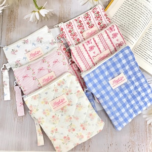 Puede incluir: Una colección de pequeños estuches con cremallera en varios diseños florales y estampados. Los estuches presentan una gama de colores, incluyendo rosa, azul y blanco, con detalles como lazos, rayas y cuadros. Cada estuche tiene una pequeña etiqueta con el nombre de la marca.