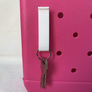 K&ouml;nnte beinhalten: Ein wei&szlig;er Schl&uuml;sselanh&auml;nger mit einem silbernen Schl&uuml;sselring und Schl&uuml;sseln, die an einer leuchtend rosa Tragetasche h&auml;ngen. Die Tasche hat mehrere runde L&ouml;cher. Der Schl&uuml;sselanh&auml;nger besteht aus einem glatten, haltbaren Material.