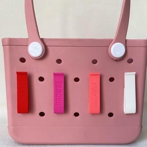 K&ouml;nnte beinhalten: Eine rosa Tragetasche mit vier farbigen Namensschildern. Die Etiketten sind rot, fuchsia, korallenfarben und wei&szlig;, mit den Namen "Tiffany", "Browne" und "Susanna" vertikal aufgedruckt. Die Tasche hat wei&szlig;e Griffbefestigungen.