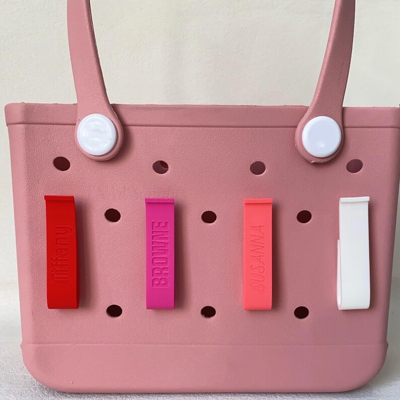 K&ouml;nnte beinhalten: Eine rosa Tragetasche mit vier farbigen Namensschildern. Die Etiketten sind rot, fuchsia, korallenfarben und wei&szlig;, mit den Namen "Tiffany", "Browne" und "Susanna" vertikal aufgedruckt. Die Tasche hat wei&szlig;e Griffbefestigungen.