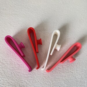 K&ouml;nnte beinhalten: Vier Kunststoffclips in Pink-, Rot- und Wei&szlig;t&ouml;nen. Jeder Clip hat ein gebogenes Design mit einem kleinen, zylindrischen Pfosten. Die Clips sind auf einer hellgrauen Oberfl&auml;che angeordnet.