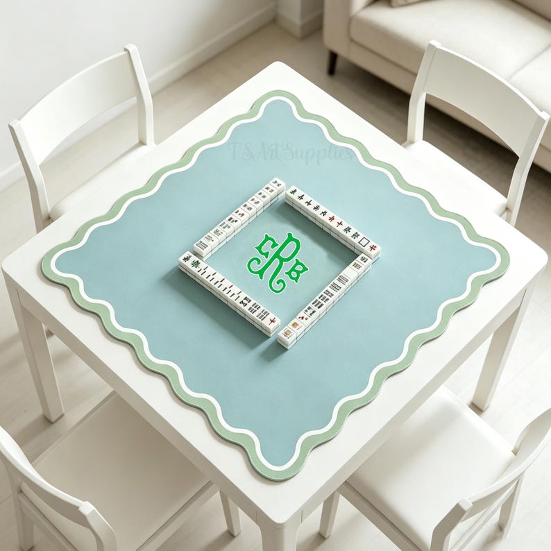 May include: A square, white table with four matching chairs. The table has a light blue mat with a scalloped green border. A set of Mahjong tiles are arranged in a square, with a green monogram in the center.