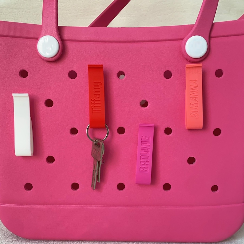 K&ouml;nnte beinhalten: Eine leuchtend pinke Tragetasche mit wei&szlig;en Akzenten und mehreren farbigen Schl&uuml;sselanh&auml;ngern. Die Schl&uuml;sselanh&auml;nger sind rot, pink und orange, mit aufgedruckten Namen. Ein Schl&uuml;sselbund h&auml;ngt am roten Schl&uuml;sselanh&auml;nger.