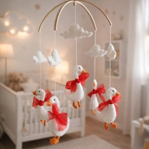 May include: A baby mobile featuring white geese with orange feet and beaks, adorned with red ribbons. The geese hang below fluffy white clouds from a wooden arch. The mobile is suspended in a nursery setting.