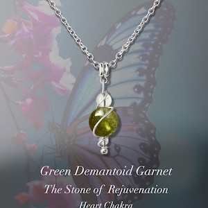 May include: A silver necklace featuring a green demantoid garnet pendant. The pendant includes a large, round, olive-green gemstone, a clear crystal bead, and a silver wire wrap. The text on the image reads "Green Demantoid Garnet, The Stone of Rejuvenation, Heart Chakra."