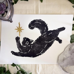May include: A black ink block print on white paper featuring a fluffy cat in a playful, leaping pose with its front paws reaching towards a gold-coloured, eight-pointed star symbol positioned above its head, creating a high-contrast artistic design.