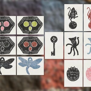 May include: A collection of twelve rectangular art prints featuring linocut-style illustrations. Designs include geometric trays with circular motifs, a key, a dragonfly, a monkey on a ball, a multi-limbed creature, and a cake with the text Welcome Home.