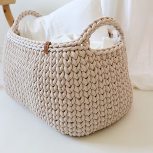 Peut inclure: Un panier de rangement en crochet beige avec deux poignées en cuir. Le panier est vide et posé sur une surface blanche.