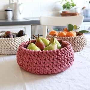May include: Three handmade crochet baskets filled with fruit. The front basket is a dusty rose color and holds green pears. The other two baskets are beige and hold plums and oranges. The baskets are on a white tablecloth.