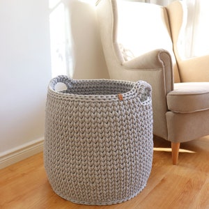 May include: A large, light gray, hand-knitted basket with two integrated handles. The basket is cylindrical and sits on a wooden floor. A light-colored armchair is in the background. The basket is approximately 15 inches in diameter and 14 inches tall.