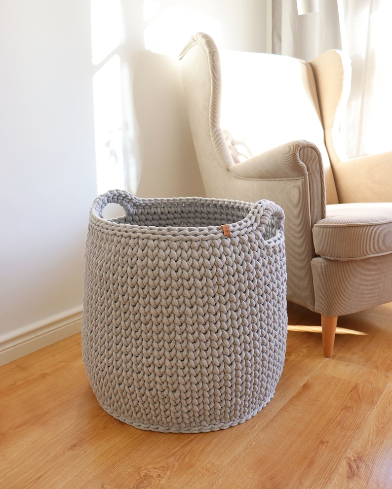 May include: A large, light gray, hand-knitted basket with two integrated handles. The basket is cylindrical and sits on a wooden floor. A light-colored armchair is in the background. The basket is approximately 15 inches in diameter and 14 inches tall.
