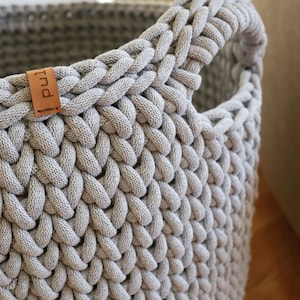May include: Close-up of a gray, woven basket with a handle. The basket is made of thick, braided material, creating a textured surface. A small, brown leather tag with the word "pul" is attached to the side. The basket is designed for storage.