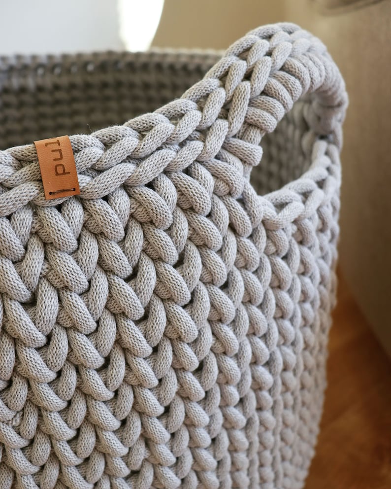 May include: Close-up of a gray, woven basket with a handle. The basket is made of thick, braided material, creating a textured surface. A small, brown leather tag with the word "pul" is attached to the side. The basket is designed for storage.