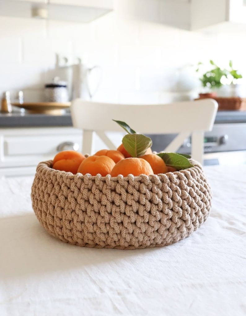 May include: A beige, crocheted basket filled with ripe apricots and green leaves sits on a white tablecloth. The basket has a thick, textured weave and a round shape. The background shows a kitchen setting.