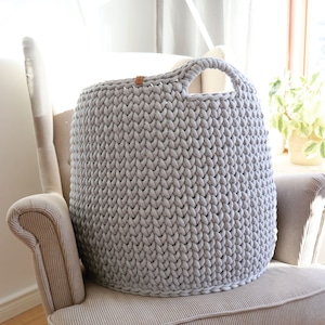 May include: A large, light gray, hand-knitted storage basket with a rounded shape and a single handle. The basket is made of thick yarn and sits on a beige armchair. A small leather tag is attached to the top of the basket.