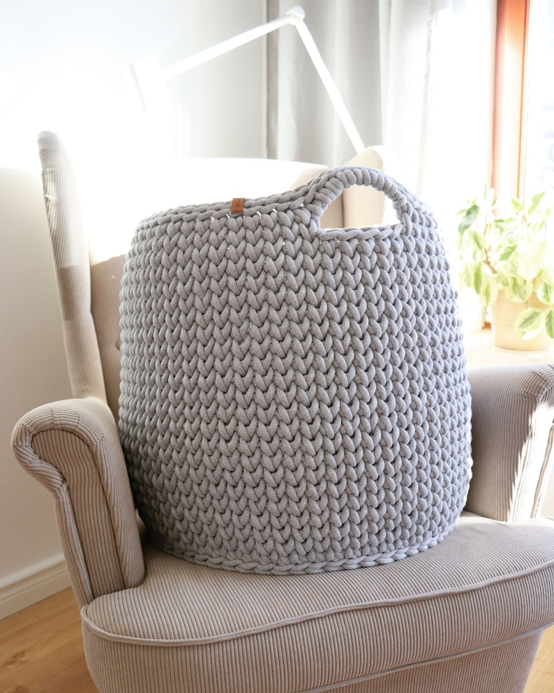May include: A large, light gray, hand-knitted storage basket with a rounded shape and a single handle. The basket is made of thick yarn and sits on a beige armchair. A small leather tag is attached to the top of the basket.