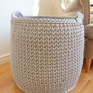 May include: A large, round, light gray knitted basket with two handles. The basket is made of thick, textured yarn and sits on a wooden floor. The basket is empty and appears to be sturdy.