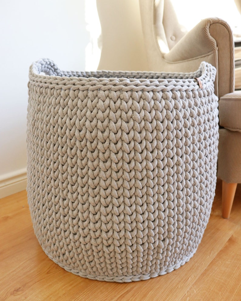 May include: A large, round, light gray knitted basket with two handles. The basket is made of thick, textured yarn and sits on a wooden floor. The basket is empty and appears to be sturdy.