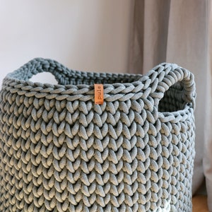 May include: A large, gray, hand-knitted storage basket with two integrated handles. The basket features a thick, textured weave and a small, brown leather tag with the word "pule" printed on it. The basket is made of a soft, durable material.