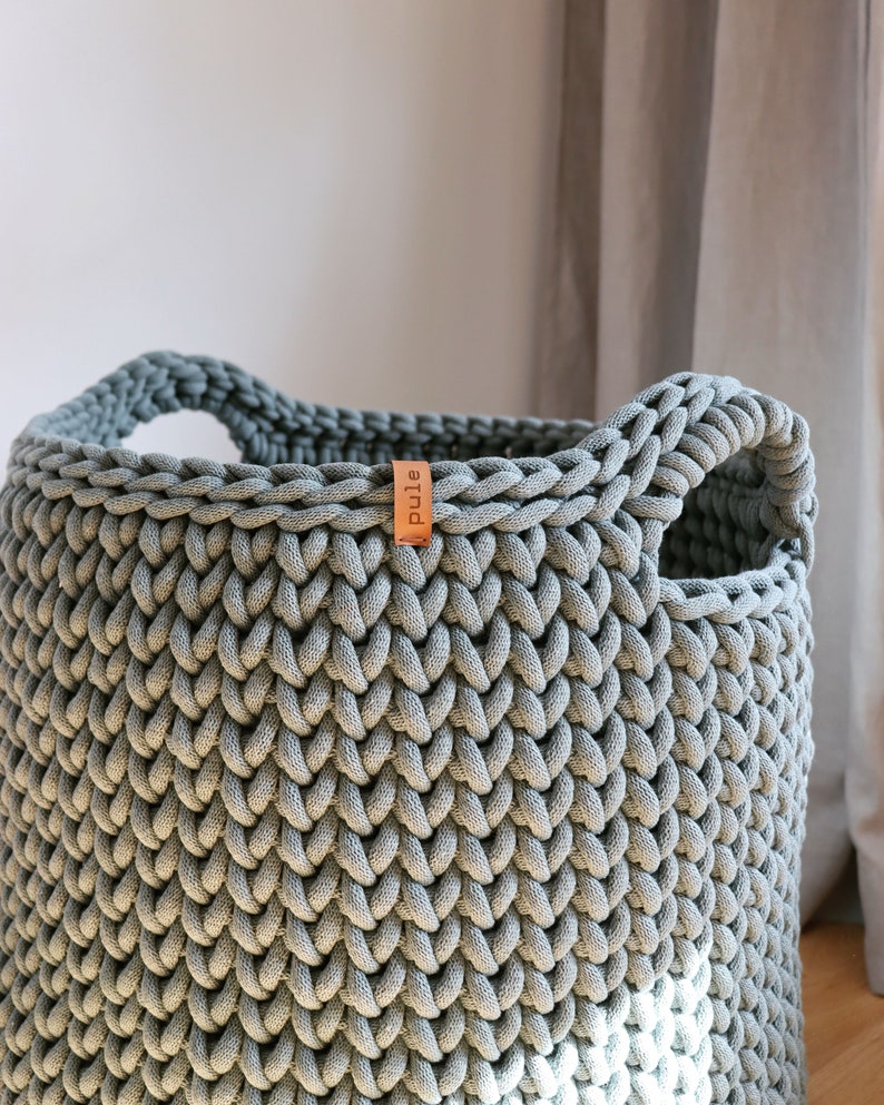 May include: A large, gray, hand-knitted storage basket with two integrated handles. The basket features a thick, textured weave and a small, brown leather tag with the word "pule" printed on it. The basket is made of a soft, durable material.
