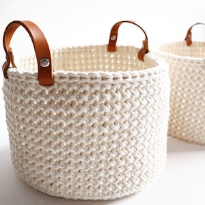 Può includere: Due cesti portaoggetti rotondi realizzati in corda intrecciata color bianco sporco con un motivo testurizzato. Ogni cesto presenta due manici in pelle marrone chiaro fissati al bordo con rivetti in metallo argentato per una presa solida.