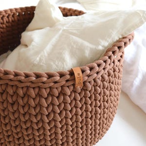 May include: A brown, hand-knitted basket with a braided rim, filled with white fabric. A small leather tag with the word "pule" is attached to the side. The basket is likely used for storage or decoration.