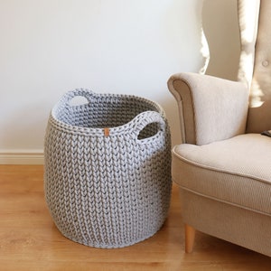 May include: A light gray, knitted storage basket with two integrated handles. The basket is cylindrical and sits on a wooden floor next to a beige armchair. A small, brown tag is attached to the basket.