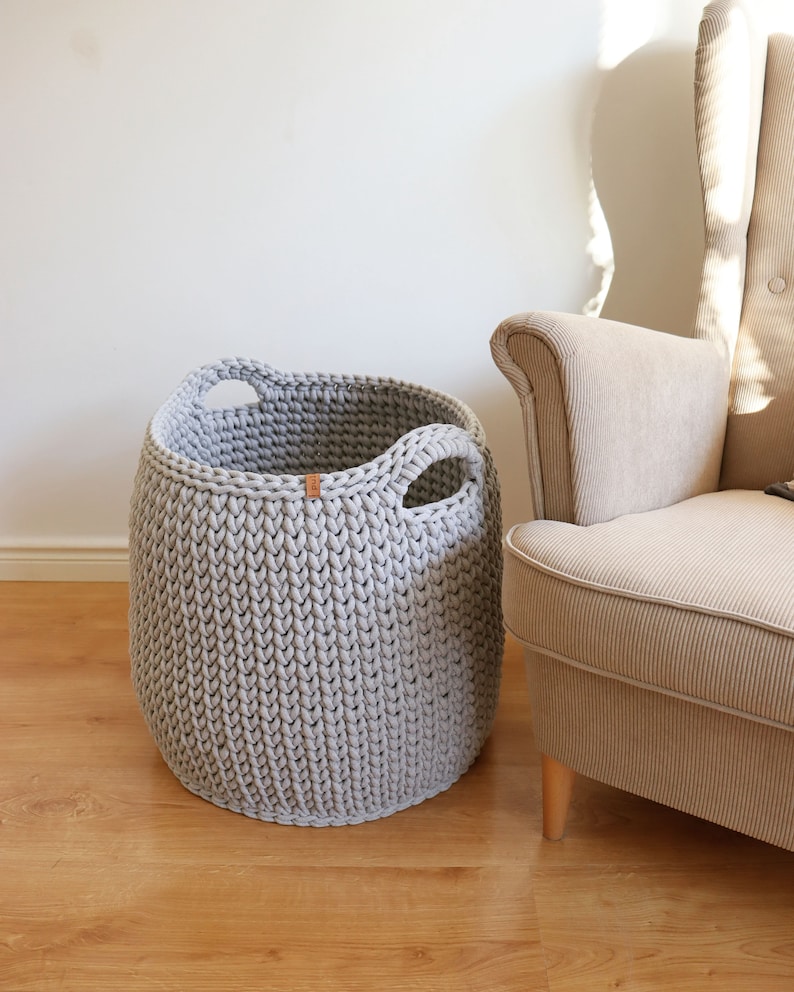 May include: A light gray, knitted storage basket with two integrated handles. The basket is cylindrical and sits on a wooden floor next to a beige armchair. A small, brown tag is attached to the basket.