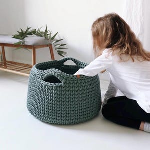 May include: A large, round, dark sage green storage basket made of thick, chunky knit yarn. The basket features two integrated handles at the top rim and a small, rectangular brown leather-like tag attached to the side of the upper edge.
