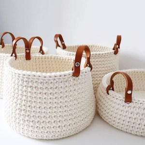 Può includere: Un set di quattro cesti portaoggetti all'uncinetto bianco sporco con manici in pelle marrone. I cesti variano per dimensioni e forma, con un design rotondo e aperto. I manici in pelle sono fissati con rivetti argentati.