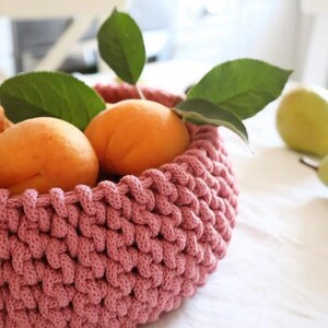 May include: A close-up of a pink crocheted basket filled with ripe apricots and green leaves. The basket is round and has a textured appearance. The apricots are orange and appear fresh. A green pear is visible in the background.