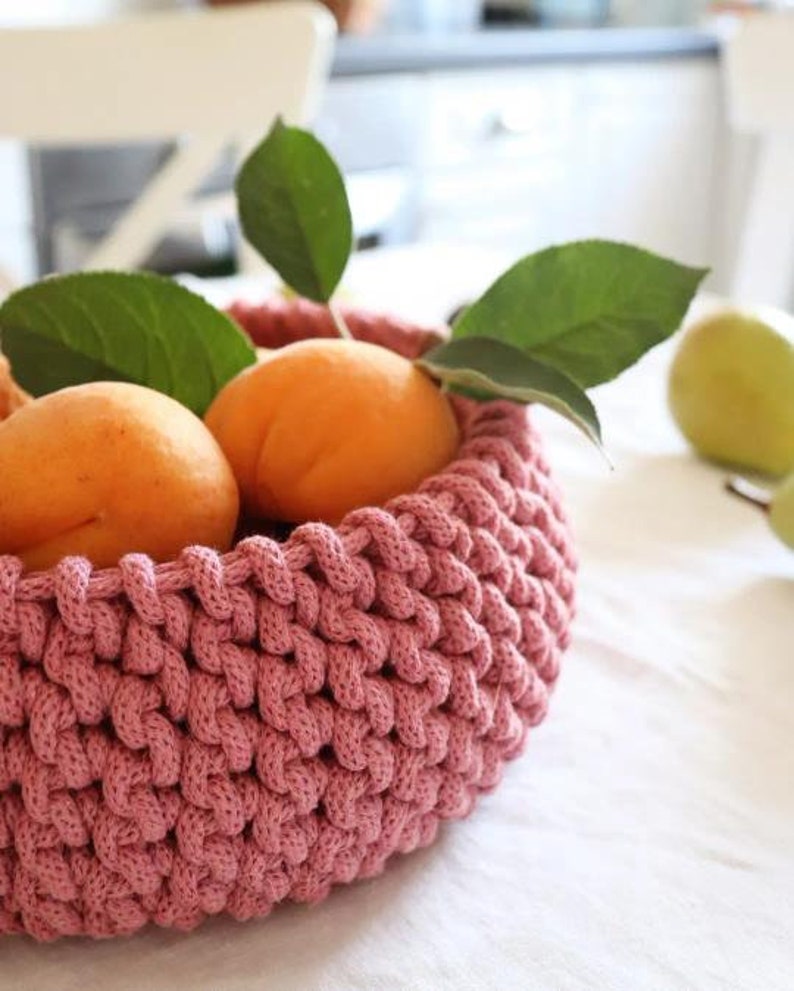May include: A close-up of a pink crocheted basket filled with ripe apricots and green leaves. The basket is round and has a textured appearance. The apricots are orange and appear fresh. A green pear is visible in the background.