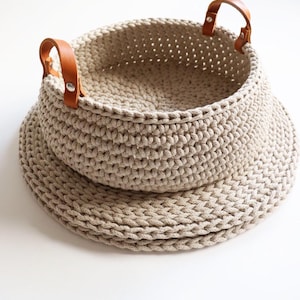 Könnte beinhalten: Ein runder, beigefarbener Häkelkorb mit einem breiten, flachen Boden und zwei hellbraunen Ledergriffen, die mit silberfarbenen Metallnieten befestigt sind. Der Korb weist ein dickes, strukturiertes Strickmuster auf.