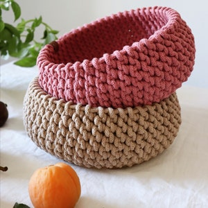 May include: Two handmade crochet baskets, one stacked on top of the other. The top basket is a dusty rose color, and the bottom basket is a light tan. The baskets are round and have a textured, woven appearance. An apricot and green leaves are in the foreground.