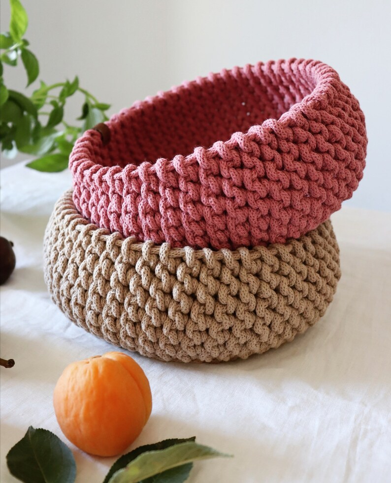 May include: Two handmade crochet baskets, one stacked on top of the other. The top basket is a dusty rose color, and the bottom basket is a light tan. The baskets are round and have a textured, woven appearance. An apricot and green leaves are in the foreground.