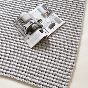 May include: A gray and white striped, hand-crocheted rug. The rug has a thick, textured appearance with horizontal stripes. An open magazine rests on the rug, displaying interior design images. The rug is on a white surface.