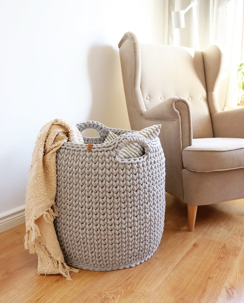 May include: A large, light gray, knitted storage basket with two handles. A beige throw blanket with fringe hangs over the side. A striped pillow is inside the basket. A beige armchair is in the background.