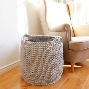 May include: A light gray, hand-knitted storage basket with two handles. The basket is cylindrical and sits on a wooden floor. A small brown tag is attached to the side. A beige armchair is in the background.