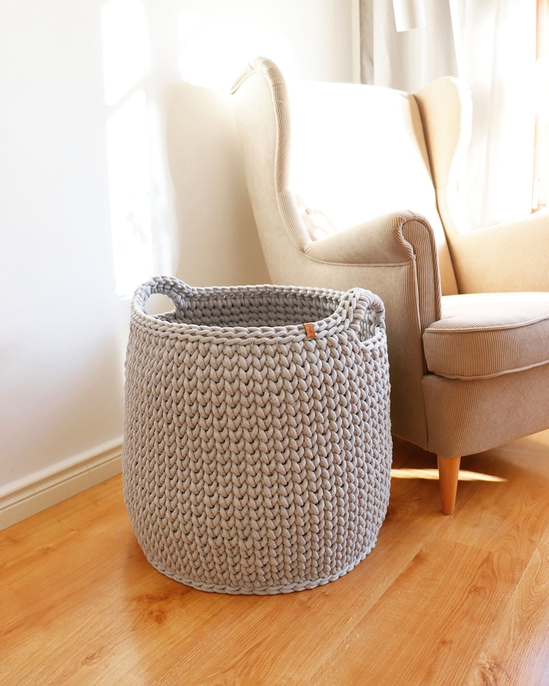 May include: A light gray, hand-knitted storage basket with two handles. The basket is cylindrical and sits on a wooden floor. A small brown tag is attached to the side. A beige armchair is in the background.