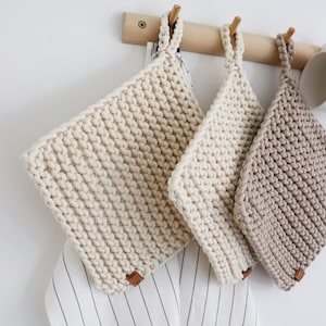 Könnte beinhalten: Drei handgefertigte, strukturierte Topflappen in Creme- und Beigetönen hängen an einer Holzstange. Jeder Topflappen hat eine Schlaufe zum Aufhängen und ein kleines braunes Lederetikett. Ein gestreiftes Handtuch ist teilweise darunter zu sehen.