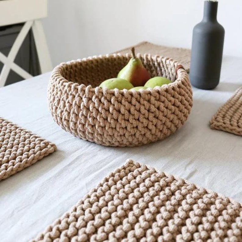 May include: A beige, hand-crocheted basket filled with green pears. The basket is round and sits on a white tablecloth, with matching coasters nearby. A dark gray bottle is in the background. The basket is approximately 10 inches in diameter.