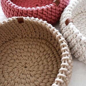 May include: Close-up of three handmade crochet baskets in varying colors. The baskets are round with a thick, textured stitch. One is a dusty rose, another is beige, and the third is speckled white. Each has a small brown leather tag.