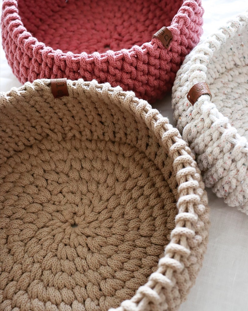 May include: Close-up of three handmade crochet baskets in varying colors. The baskets are round with a thick, textured stitch. One is a dusty rose, another is beige, and the third is speckled white. Each has a small brown leather tag.