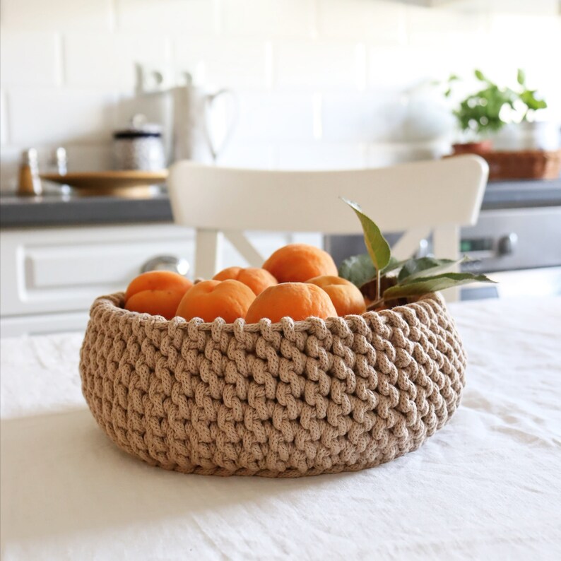 May include: A round, beige, crocheted basket filled with ripe, orange apricots. The basket sits on a white tablecloth, with a white chair and kitchen in the background. A few green leaves are tucked among the fruit.