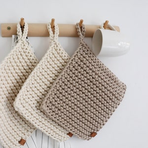 Op de afbeelding: Drie gehaakte pannenlappen in cr&egrave;me- en beigetinten hangen aan een houten rail. Elke vierkante pannenlap heeft een lus en een klein bruin label. Er hangt ook een witte mok. De pannenlappen zijn waarschijnlijk ongeveer 20 cm vierkant.