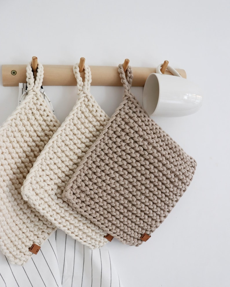Op de afbeelding: Drie gehaakte pannenlappen in cr&egrave;me- en beigetinten hangen aan een houten rail. Elke vierkante pannenlap heeft een lus en een klein bruin label. Er hangt ook een witte mok. De pannenlappen zijn waarschijnlijk ongeveer 20 cm vierkant.